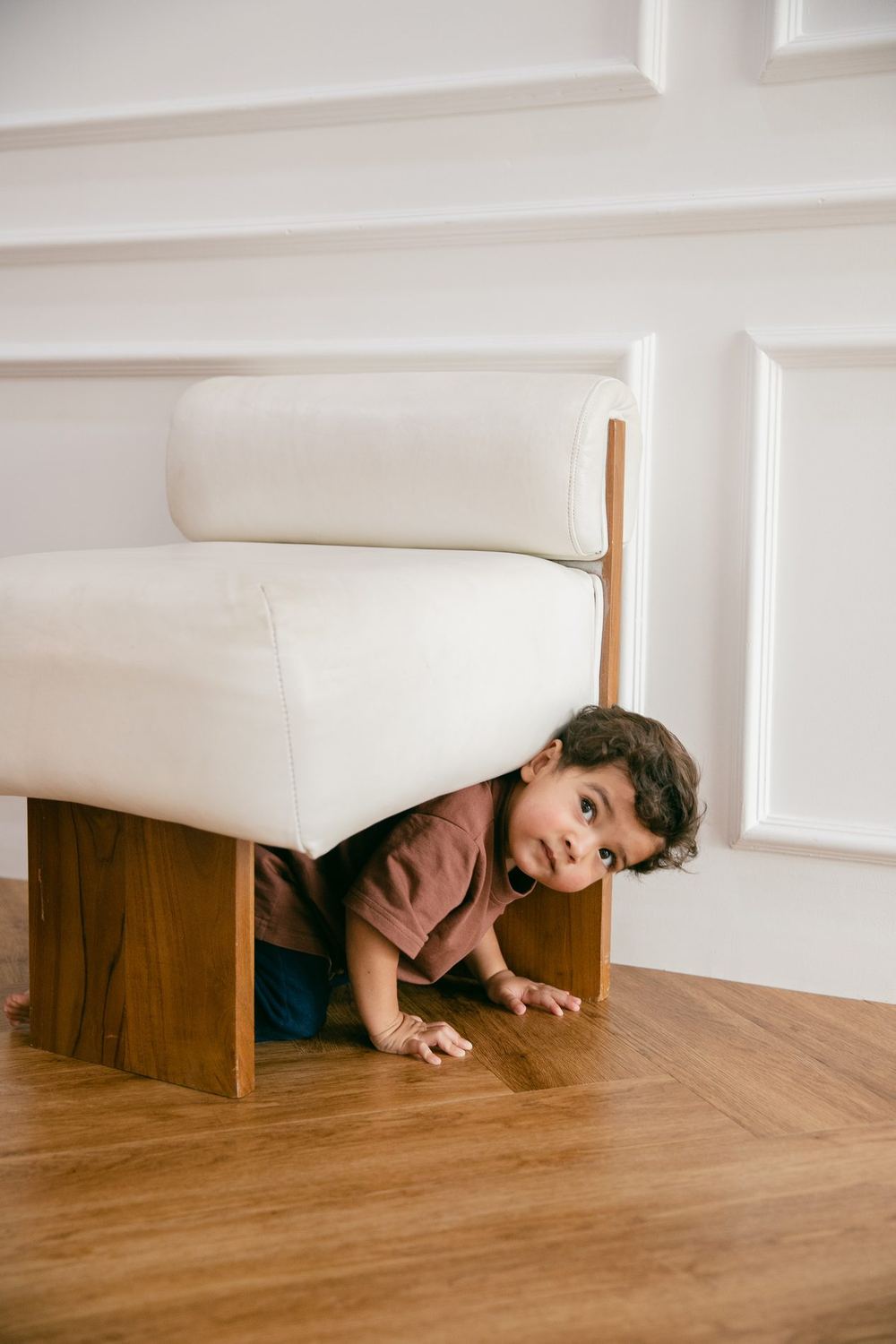 A young child peering out from underneath a sofa in a bright room